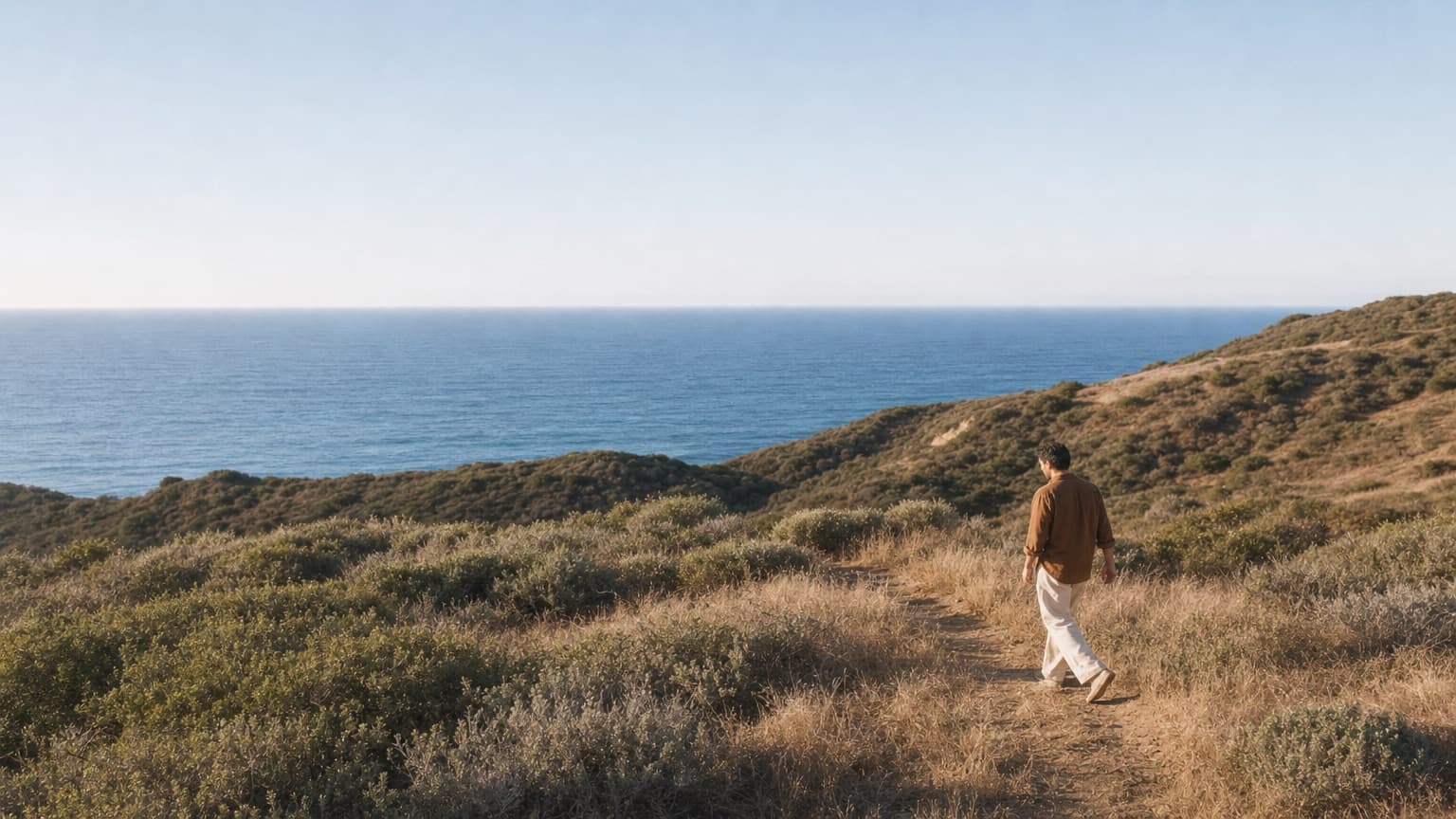 A man walking a narrow path through coastal scrub towards the sea